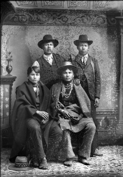 Studio portrait of two Ho-Chunk men posing sitting in front of two Ho-Chunk men posing standing in front of a painted backdrop. All are wearing hats except the man sitting on the right whose hat is on the ground at his feet. The two sitting men are also wrapped in blankets. Men identified include George Lyons Lowe standing on the right, and Jasper Blowsnake sitting on the left.