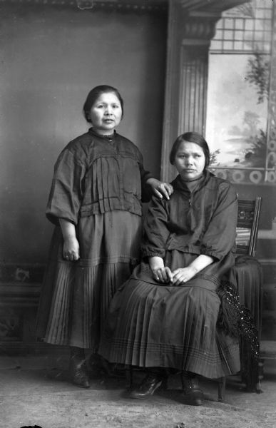 Studio portrait of two Ho-Chunk women wearing contemporary dark dresses and short hair posing in front of a painted backdrop. The woman on the left, identified as Connie Blackhawk, is standing and the woman on the right, identified as Elizabeth Thundercloud, is sitting.