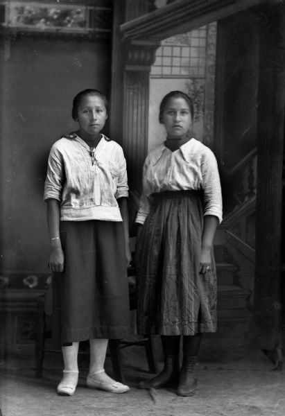Studio portrait of two young Ho-Chunk women posing standing in front of a painted backdrop. They are wearing contemporary dark skirts and light blouses. Identified as Mary Lewis on the left and Hazel Thunder on the right.
