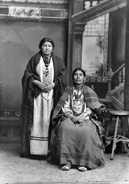 Studio portrait of two Ho-Chunk women posing in front of a painted backdrop. The woman on the left is standing and wearing a shawl, several necklaces and rings, and the woman on the right is sitting and wearing a shawl and several necklaces, file bracelets, and earrings. Identified as White Oaktree standing on the left, and the sister of John Johnson sitting on the right.