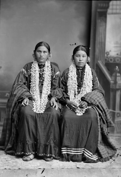 Studio portrait of two young Ho-Chunk women posing sitting in front of a painted backdrop. They are both wearing several necklaces and earrings, and are holding a plaid shawl over their outside arm. Identified as the wife of Frank Lincoln on the left, and the wife (Stand Straight) of Jasper Blowsnake on the right.