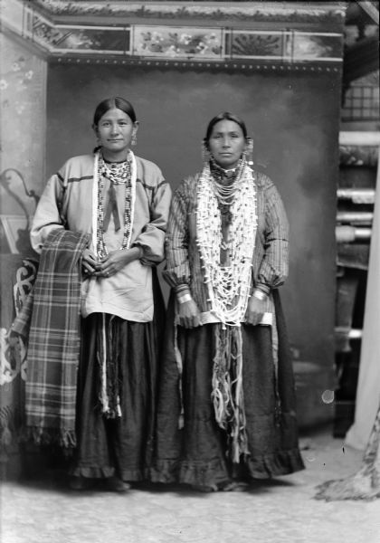 Studio portrait of two Ho-Chunk women posing standing in front of a painted backdrop. They are both wearing regalia, including dark skirts and light-colored blouses, several necklaces, and several earrings. The woman on the left, identified as Blanche Blackhawk, is holding a plaid shawl. The woman on the right, identified as Ella Bill/Bell, has more accessories and file bracelets.