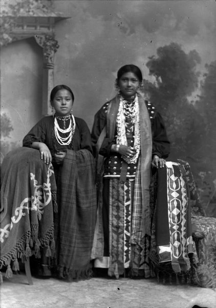 Studio portrait of two Ho-Chunk girls posing standing in front of a painted backdrop. They are wearing regalia, including several necklaces, earrings, and file bracelets. The girl on the left, identified as Annie Standing Water, is leaning on a stuffed chair and is holding a plaid shawl. The older girl on the right, identified as Bleeds the Bear, has her hand on a chair draped with a geometric shawl.