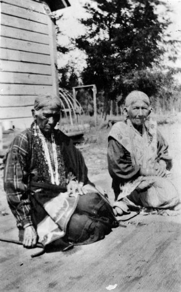 Copy photograph of two elderly Ho-Chunk women posing sitting on the ground in front of a building with a lodge frame in the background. Identified as Strikes A Tree on the left, and The Small One on the right. Reportedly they were the last ones to live in traditional lodges.