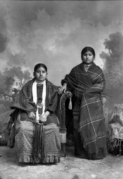 Studio portrait of two young Ho-Chunk women posing in front of a painted backdrop. The woman on the left, Identified as Red Bird, the wife of Jesse Brown, is sitting and wearing several necklaces and earrings. The woman on the right, identified as the second wife of George Lowe is standing and wrapped in a shawl wearing several earrings.