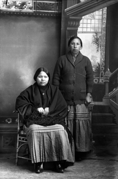 Studio portrait of two Ho-Chunk women posing in front of a painted backdrop. The woman on the left, identified as Connie Blackhawk, is sitting and wearing a fringed shawl. The woman on the right, identified as Mary Falcon Whitewater, the wife of Homer Snake, is standing and wearing a button-down sweater.