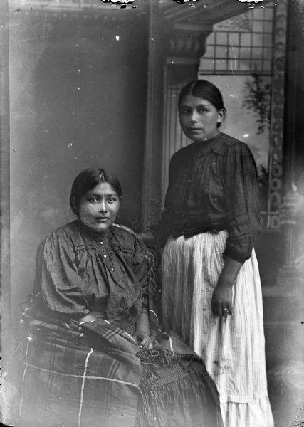 Studio portrait of two young Ho-Chunk women posing in front of a painted backdrop. They are both wearing contemporary dress. The woman on the left, identified as Edna Grizzly Bear, is sitting and wearing a dark dress and shawl. The woman on the right, identified as Nellie Wallace Black Deer, is wearing a light skirt and dark blouse and is standing with her hand on the other woman's shoulder.