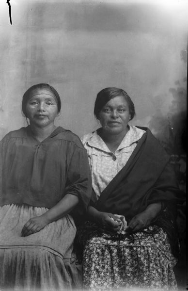 Studio portrait of two Ho-Chunk women posing sitting and wearing contemporary clothes in front of a painted backdrop. The woman on the left is identified as Lucy White Davis. The woman on the right, identified as Jennie Young Thunder, is also wearing a fringed shawl.