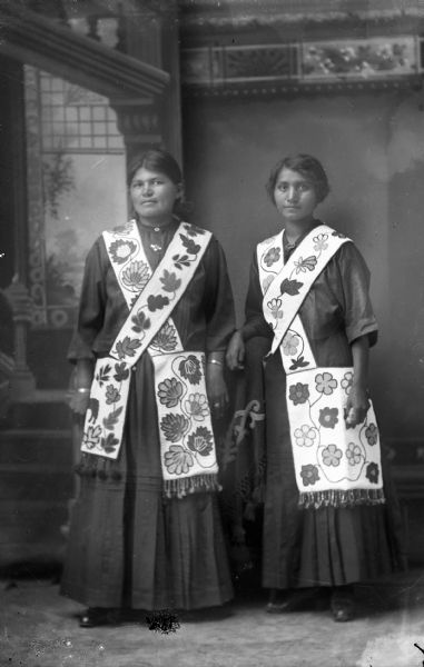 Studio portrait of two Ho-Chunk women posing standing in front of a painted backdrop. They are wearing contemporary dress except for the woodland-style Chippewa bandoleers on each. Identified as Flora Bear Heart on the left, and Ethel Windblowe on the right.