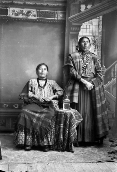 Studio portrait of two elderly Ho-Chunk women posing in front of a painted backdrop. The woman on the left is sitting and wearing several short necklaces, and holding a plaid shawl/blanket on her lap. The woman on the right is standing, wearing several necklaces, and holding a striped shawl/blanket, with another plaid shawl/blanket over her right shoulder. A liquor bottle is sitting on a small table draped with a plaid cloth in between them. Identified as White Oak Tree, the mother of George Johnson, on the left, and the wife of wife of King of Thunder George Lowe and mother of Gilbert Lowe standing on the right.