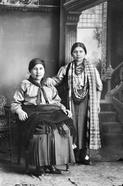 Studio portrait of two Ho-Chunk women posing in front of a studio backdrop. The woman sitting on the left is wearing contemporary clothing, with a fringed shawl over her lap, and is identified as Elizabeth Thundercloud Hall. The woman standing on the right is wearing several long necklaces, and a shawl draped over her shoulder, and is identified as Kate Miner.