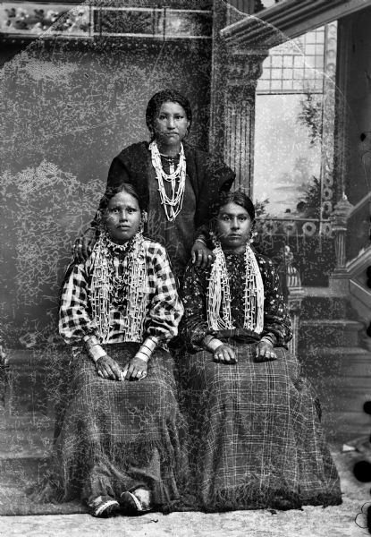 Studio portrait of three young Ho-Chunk women posing in front of a painted backdrop. Two of the women are sitting in front of another woman standing. They are all wearing several necklaces and earrings, in addition to contemporary clothing. The seated women have shawls/blankets over their laps, and the standing woman has a shawl/blanket over her shoulders.