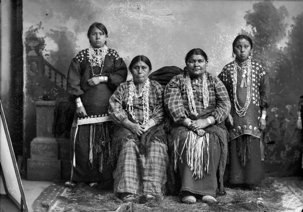 Studio portrait of two Ho-Chunk women posing sitting between two Ho-Chunk women posing standing in front of a painted backdrop. They are all wearing regalia, including beaded moccasins, bead cloth skirts, fingered sashes, several necklaces, and earrings. Identified from left to right as: a Climer daughter, wife of ? Climer, wife of Little Creek, and another Climer daughter.