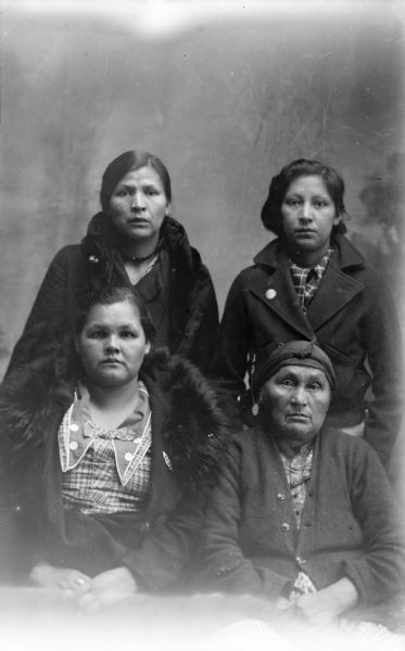 Studio portrait of two Ho-Chunk women posing sitting in front of two Ho-Chunk women posing standing. They are all wearing contemporary clothing, and three of the women are wearing winter coats. The women standing are identified as Dora Decorah on the left, and Helen Lincoln on the right. The women sitting in the front are identified as Mabel Kingsley Lincoln on the left, and Mrs. Lincoln on the right.