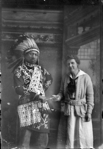 Studio portrait of a Ho-Chunk man posing standing and wearing full regalia, including a Sioux headdress, bandoleers, and bead work clothing. He is holding a calumet pipe which he is handing to a white woman in contemporary clothing. They are both standing in front of a painted backdrop. Identified as Thomas Thunder and Rachel Commons or Francis Densmore, an anthropologist and the daughter of Dr. John Commons of the University of Wisconsin.