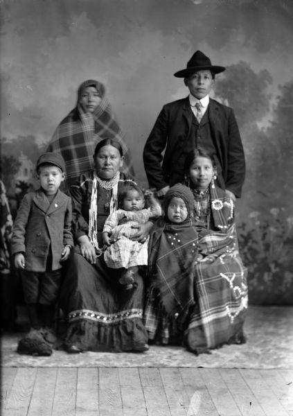 Studio group portrait in front of a painted backdrop. Two Ho-Chunk women are wearing several necklaces and earrings, and three Ho-Chunk children posing sitting and standing in front of a Ho-Chunk man wearing a suit, necktie, and hat, and a Ho-Chunk woman wrapped in a shawl. The man standing is identified as William Miner, and the woman sitting on the left is his wife. The woman sitting on the right is Kate Miner.