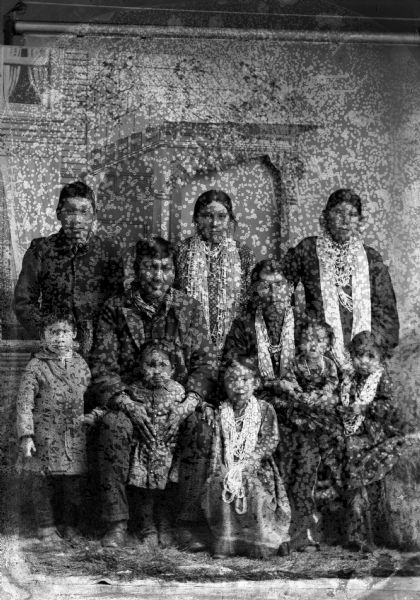 Studio portrait in front of a painted backdrop of a Ho-Chunk man, woman, and five children posing sitting and standing in front of two Ho-Chunk girls and a Ho-Chunk boy. The female individuals are wearing several necklaces and earrings, and the male individuals are wearing suits. The man sitting is identified as King of Thunder, and the boy standing on the left is David Decorah.