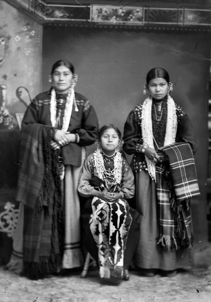 Studio portrait in front of a painted backdrop of a Ho-Chunk girl posing sitting on a stool and wearing ribbon work skirt, between two Ho-Chunk women posing standing and holding shawl/blankets over their outside arms. All the women are wearing regalia including several necklaces, earrings, and file bracelets. The woman on the left is Blanche Blackhawk, and the woman standing on the right is Mabel White.
