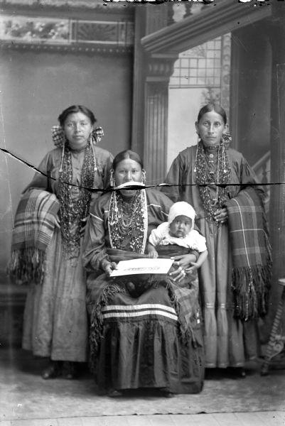 Studio portrait in front of a painted backdrop of a Ho-Chunk woman posing sitting with a shawl across her lap, and holding an infant and a photograph. Standing behind on the left and right are two Ho-Chunk women posing standing and holding shawls/blankets over their outside arms. All the women are wearing several necklaces and earrings. Identified, from left to right, as Lucy Davis, Mary Decorah, and Ruth Honka, the wife of Andrew Blackhawk.