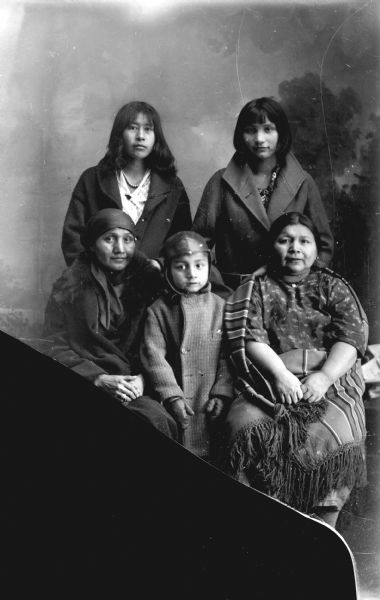 Studio portrait in front of a painted backdrop of two Ho-Chunk women posing sitting, and a Ho-Chunk boy posing standing, in front of two Ho-Chunk women posing standing. All of them are wearing contemporary dress. Identified in front, from left to right, as Elizabeth Thundercloud, Myron Funmaker, and, wrapped in a shawl, Flora Thundercloud-Funmaker-Bear Heart. Standing behind on the left is Ruth Greengrass and on the right is Jesse (Tebo) Grayhair.