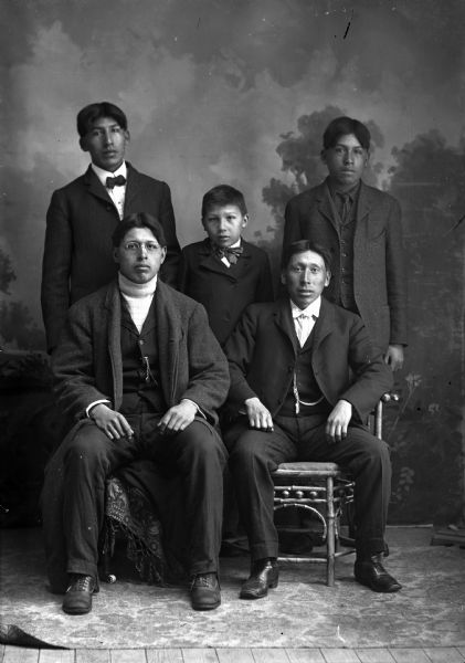 Studio portrait in front of a painted backdrop of two young Ho-Chunk men posing sitting in front of two young Ho-Chunk men flanking a Ho-Chunk boy. All of them are wearing suits and neckties or bow ties, except the man sitting on the left. Identified as, sitting from left to right: Fred Kingswan and Theodore Lowe; standing: Gilbert Lowe, boy?, and Theodore Lowe.