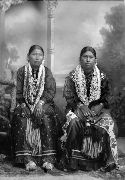 Studio portrait of two Ho-Chunk women posing sitting in front of a painted backdrop near a prop wood fence. They are wearing regalia, including several bugle and shell necklaces, rings, file bracelets, trader ear bob earrings, etc. Identified as the wife of Ulysses White Bear on the left, and the wife of White Bear on the right.