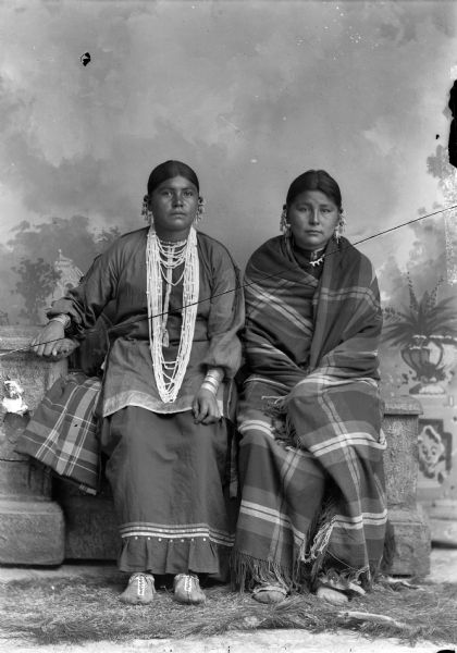 Studio portrait of two young Ho-Chunk women posing sitting in front of a painted backdrop on a prop stone wall. They are both wearing several earrings. The woman on the left is wearing several necklaces and the woman on the right is wrapped in a shawl. Identified as the wife of John Davis on the left and the sister of John Davis on the right.