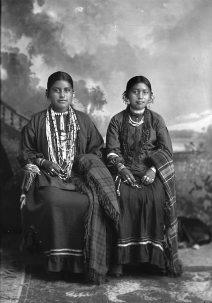Studio portrait in front of a painted backdrop of two young Ho-Chunk women posing sitting. They are both wearing several necklaces, file bracelets, rings, earrings, and shawls draped over their left arms. Identified as Ida Lizzie Decorra, Blowsnake, Bearheart (Woo Roo Shiek E Skae Win Kah) (Real Wampum Woman) on the left and the wife of Yellow Thunder on the right. 