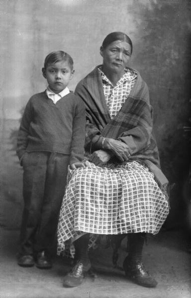 Studio portrait  in front of a painted backdrop of a Ho-Chunk boy standing and leaning against a Ho-Chunk woman sitting. The woman is wearing a shawl wrapped around her shoulders. Identified as Mary White-Swan, the wife of Frank Swan, and her son, Henry Swan.