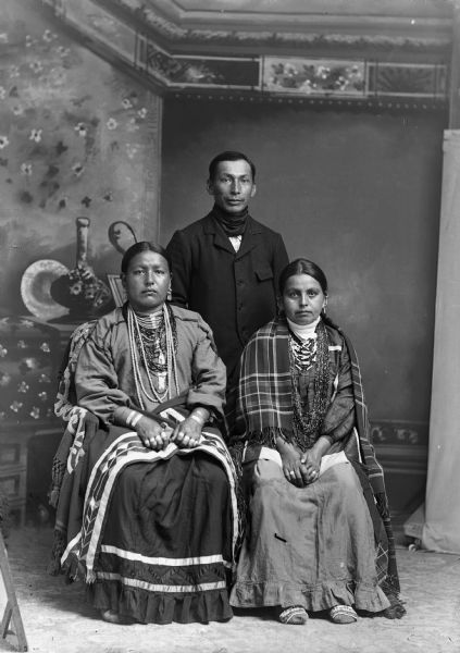 Studio portrait of two Ho-Chunk women posing sitting in front of a Ho-Chunk man standing in front of a painted backdrop. The women are wearing several necklaces, earrings, rings, and file bracelets. The woman on the left has a shawl wrapped around her waist and the woman on the right has a shawl wrapped around her shoulders. The Ho-Chunk man, John Stacy, is wearing a suit and a bandana around his neck.