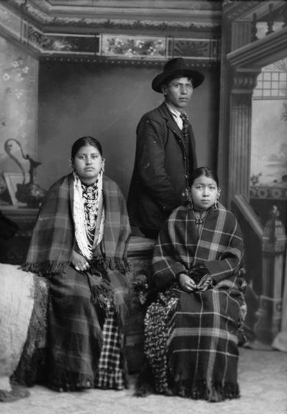 Studio portrait  of two young Ho-Chunk women wearing several necklaces, earrings, and shawls, sitting in front of a Ho-Chunk man posing standing and wearing a suit, necktie, and hat. Standing, Frank C. Thunder.  Sitting (l to r) Ida Lizzie Decorra, Blowsnake, Bearheart ([Woo Roo Shiek E Skae Win Kah] Real Wampum Woman) and name illegible. In the background is a painted backdrop.