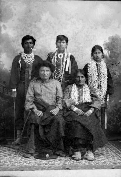 Studio portrait in front of a painted backdrop of a middle-aged Ho-Chunk man and woman posing sitting with shawls wrapped around their waists. They are in front of two young Ho-Chunk men and a young Ho-Chunk woman posing standing. The women are wearing several necklaces and earrings, and the young men are wearing necklaces and streamers. The young man standing on the left has a pipe in his mouth, and the older man is wearing a hat, while another hat is on the ground by his feet. Identified from left to right, standing Moheek Thunder Cloud, unidentified, and the daughter of Four Cloud, and sitting is Four Cloud and his wife.