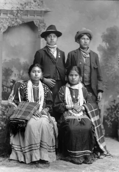 Studio portrait in front of a painted backdrop of two young Ho-Chunk women posing sitting and wearing several necklaces, earrings, file bracelets, and rings, and with shawls over their arms. They are in front of two young Ho-Chunk men posing standing. They are wearing suits and hats. Names, sitting (l to r), unknown and Emma Thunder, Littlesoldier (We Pa Ma Ka Ra Win Kah), and standing (l to r) Johnnie Thunder and John Rainbow (Nebraskan Winnebago).