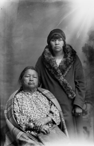 Studio portrait in front of a painted backdrop of a Ho-Chunk woman posing sitting on the left wearing with a plaid shawl over her shoulders, and a young Ho-Chunk woman posing standing on the right wearing a fur collar coat and hat, circa 1910-20s.