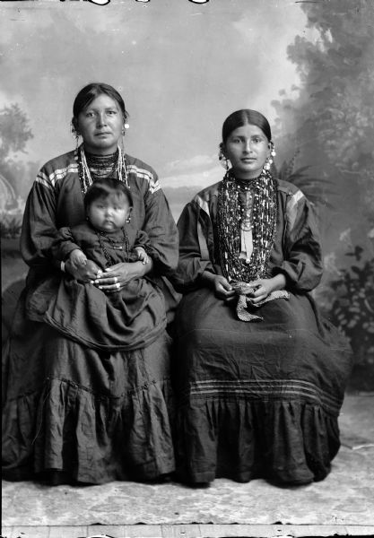 Studio portrait in front of a painted backdrop of two Ho-Chunk women posing sitting and wearing several necklaces, earrings, and rings. The woman on the left is also holding a small Ho-Chunk girl on her lap.