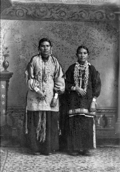 Studio portrait in front of a painted backdrop of two Ho-Chunk women posing standing and wearing several necklaces, earrings, and file bracelets. They both have their right hands holding the bottom of their necklaces.