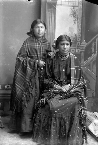 Studio portrait in front of a painted backdrop of a Ho-Chunk girl posing standing on the left wrapped in a plaid shawl, and a young Ho-Chunk woman posing sitting on the right, wearing several short necklaces, file bracelets, rings, and a shawl over her left shoulder. They are both wearing contemporary clothing.
