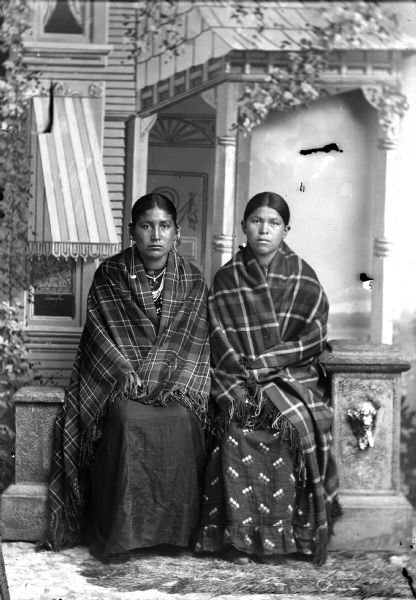 Studio portrait of two young Ho-Chunk women posing sitting on a prop stone wall in front of a painted backdrop. They are wearing plaid shawls over their shoulders and are wearing earrings. The woman on the left is also wearing necklaces.