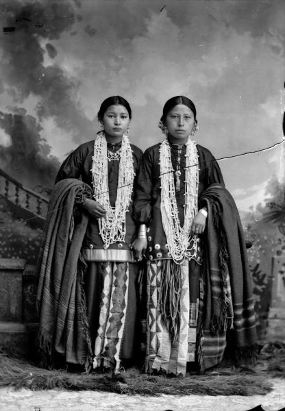 Studio portrait in front of a painted backdrop of two young Ho-Chunk women posing standing. They are holding hands and wearing several necklaces, earrings, file bracelets, and ribbon-work, and are holding shawls draped over their outside arms.