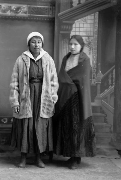 Studio portrait in front of a painted backdrop of two young Ho-Chunk women posing standing. The woman on the left is wearing a cap, and a long button-down sweater over a dress. The woman on the right is wrapped in a dark-colored fringed shawl.