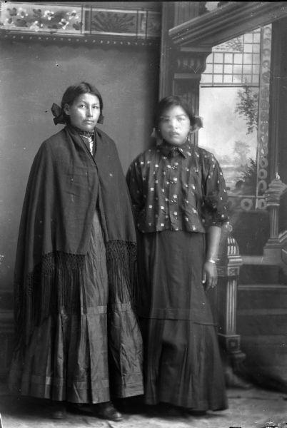 Studio portrait in front of a painted backdrop of two young Ho-Chunk women posing standing. The woman on the left is wrapped in a dark-colored fringed shawl, and the woman on the right is wearing a printed blouse, with her face obscured from movement.