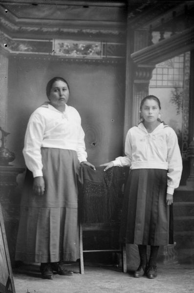 Studio portrait in front of a painted backdrop of two young Ho-Chunk women posing standing. They are flanking a chair in the center which is draped with a dark-colored fringed shawl. Both women are wearing light-colored blouses and dark-colored skirts.