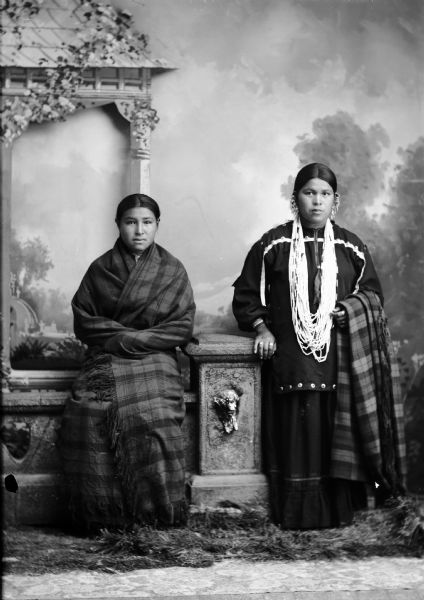 Studio portrait in front of a painted backdrop of a young Ho-Chunk woman posing sitting on a prop stone wall, wrapped in a plaid fringed shawl on the left. A young Ho-Chunk woman is posing standing, wearing several necklaces and earrings, and holding a plaid fringed shawl draped over her left arm, on the right.