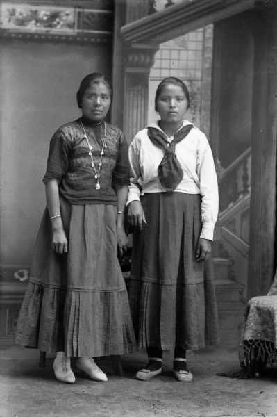 Studio portrait in front of a painted backdrop of two young Ho-Chunk women posing standing. They are wearing short hairstyles and contemporary clothing. The woman on the left is also wearing a Winnebago chain and coin necklace, and bracelets.