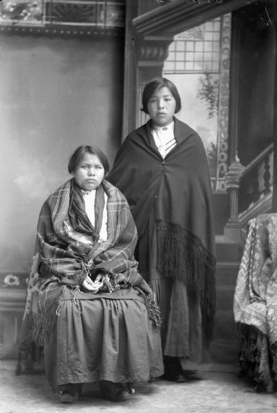 Studio portrait in front of a painted backdrop of a Ho-Chunk woman posing sitting on the left wrapped in a plaid fringed shawl, and a Ho-Chunk woman posing standing on the right wearing a fringed shawl. Both women are wearing short hairstyles.