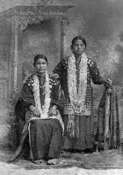 Studio portrait in front of a painted backdrop of two Ho-Chunk women. The woman on the left is posing sitting, and the other Ho-Chunk woman is posing standing on the right, with her right hand on the left shoulder of the woman sitting, and her left hand on a prop wooden fence draped with a shawl. Both woman are wearing regalia in the form of several necklaces, earrings, file bracelets, and brooch blouses.