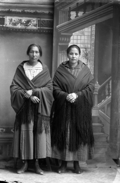 Studio portrait in front of a painted backdrop of two Ho-Chunk women posing standing. They are wrapped in fringed shawls over contemporary clothing.