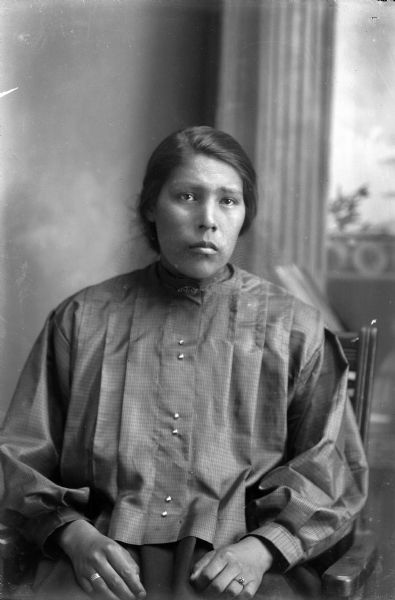 Studio portrait in front of a painted backdrop of a young Ho-Chunk woman posing sitting in a chair, wearing a contemporary blouse and two rings.