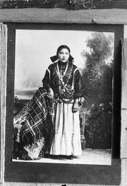 Copy photograph of a studio full-length portrait in front of a painted backdrop of a young Ho-Chunk woman posing standing with her right arm resting on a chair covered with a cloth. She is wearing several bead bandoleers, necklaces, earrings, and rings.