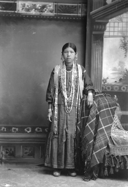 Studio portrait in front of a painted backdrop of a young Ho-Chunk woman posing standing. She has her left arm on a chair which is draped with a shawl. She is wearing several necklaces, earrings, streamers, file bracelets, and earrings.